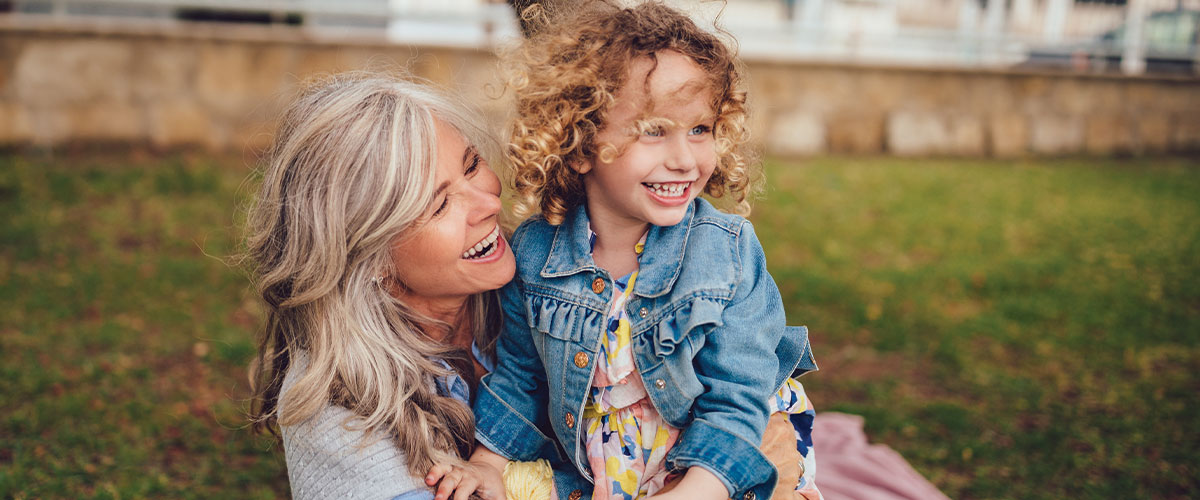 An older woman smiles warmly at a young child sitting beside her in a grassy outdoor setting, capturing a joyful moment of connection and shared happiness.